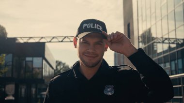 happy young policeman looking at camera and adjusting cap outdoors  clipart