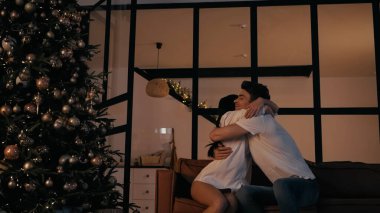 Genç çift Noel ağacının yanındaki kanepede birbirine sarılıyor.