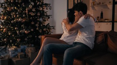 Genç çift Noel ağacının yanındaki kanepede birbirlerine hediyelerle sarılıyorlar.