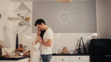 Modern mutfakta beyaz gömlekli esmer kadına sarılan memnun bir adam. 