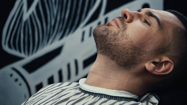 Bearded man in hairdressing cape sitting in barbershop 