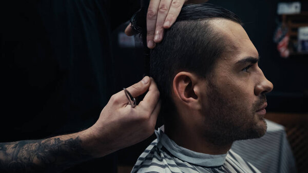 Hairstylist holding scissors and combing hair of man in hairdressing cape in salon 