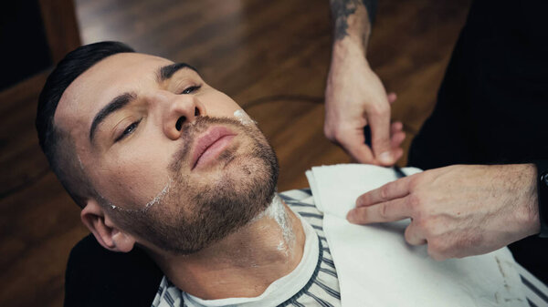 High angle view of barber standing near customer with foam on beard in barbershop 