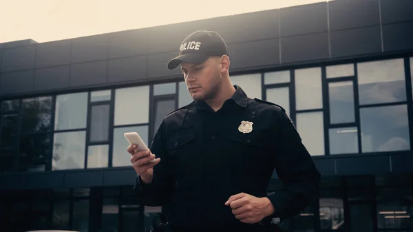 policeman in uniform and badge using smartphone on urban street