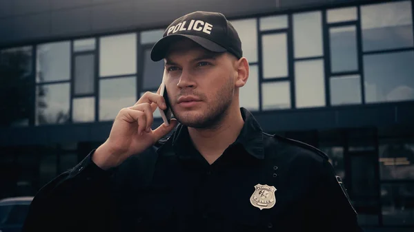 serious policeman in uniform and cap talking on mobile phone on urban street