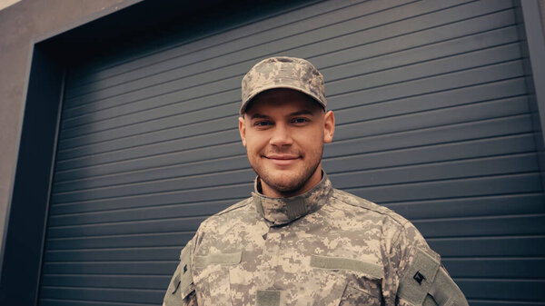 cheerful young soldier in uniform and cap smiling while looking at camera near building 