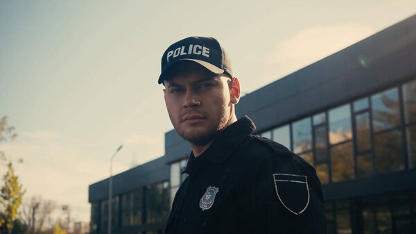 serious policeman looking at camera while standing in cap and uniform near building 