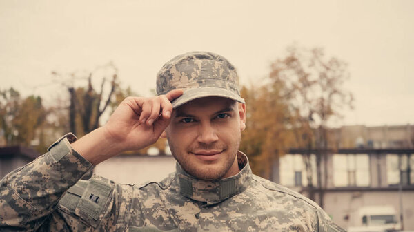 Smiling soldier in uniform adjusting cap and looking at camera outdoors 