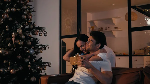 happy woman hugging boyfriend with present sitting on couch near christmas tree