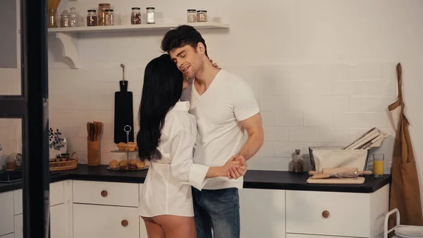 smiling man hugging woman in shirt while dancing in modern kitchen