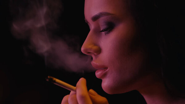 Young woman holding cigarette near smoke on black background 