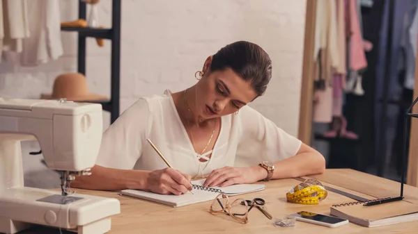 Seamstress writing on notebook near tape measure and sewing machine in workshop