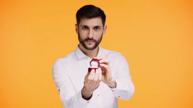 Beyaz tişörtlü sakallı bir adam nişan yüzüğüyle kutusunu tutarken evlenme teklifini sarıda izole ediyor.