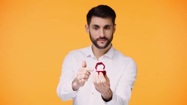 Beyaz tişörtlü sakallı bir adam nişan yüzüğüyle kutuyu gösterirken evlenme teklifini sarıda izole ediyor.