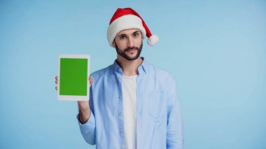Kırmızı Noel Baba şapkalı sakallı adam elinde yeşil ekranlı dijital tablet var. 