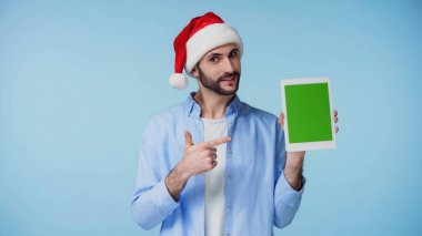 Kırmızı Noel Baba şapkalı mutlu adam mavi ekranlı dijital tableti işaret ediyor. 