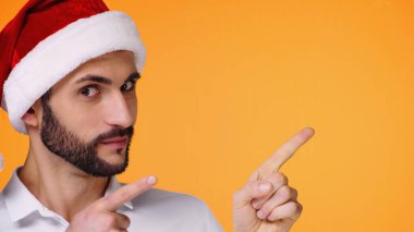 Kırmızı Noel Baba şapkalı sakallı adam parmaklarıyla işaret ediyor ve sarı renkli kameraya bakıyor.