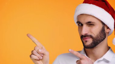 Kırmızı Noel Baba şapkalı sakallı adam sarıda izole edilmiş göz kırparken parmaklarıyla işaret ediyor.