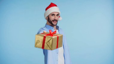 Kırmızı Noel Baba şapkalı sakallı adam mavi renkte paketlenmiş Noel hediyesi veriyor.
