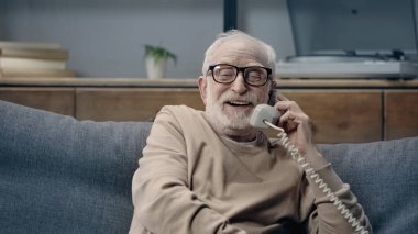Gözlüklü neşeli son sınıf öğrencisi klasik telefonla konuşuyor ve evdeki kanepede oturuyor.