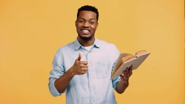 Mavi gömlekli mutlu Afro-Amerikan adam başparmağını kaldırıyor ve sarı üzerine izole bir kitap tutuyor.