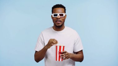 Odaklanmış Afro-Amerikan gözlüklü adam film izliyor ve mavi üzerine izole edilmiş patlamış mısır tutuyor.