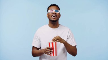3D gözlüklü mutlu Afrikalı Amerikalı adam film izliyor ve elinde patlamış mısır kovası tutuyor.