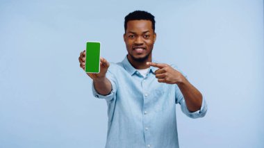 Mutlu Afro-Amerikan tişörtlü adam mavi ekranda yeşil ekranlı akıllı telefonu işaret ediyor.
