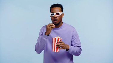 Üç boyutlu gözlüklü Afro-Amerikan bir adam film izliyor ve elinde patlamış mısır kovası tutuyor.