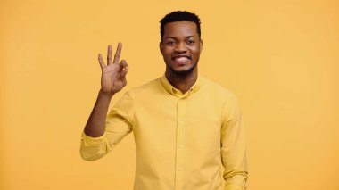 positive african american man in shirt showing ok sign isolated on yellow  clipart