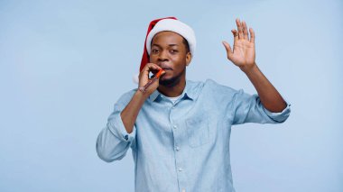 Noel Baba şapkalı Afro-Amerikan bir adam parti kornası çalıyor ve mavi üzerine izole edilmiş bir şekilde el kol hareketi yapıyor.