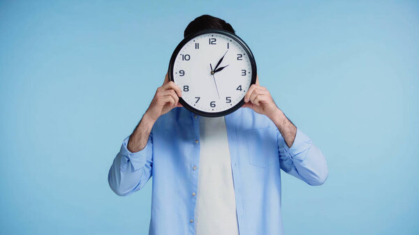 man in shirt holding clock while covering face isolated on blue background 