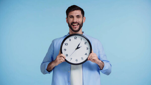 cheerful man in shirt holding clock while looking at camera isolated on blue background 