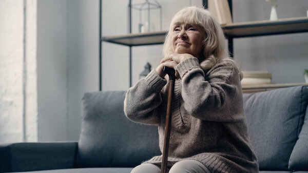 sad senior woman with dementia sitting on sofa while leaning on walking cane 