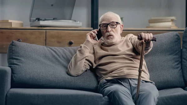 surprised senior man talking on smartphone and sitting on sofa with walking cane