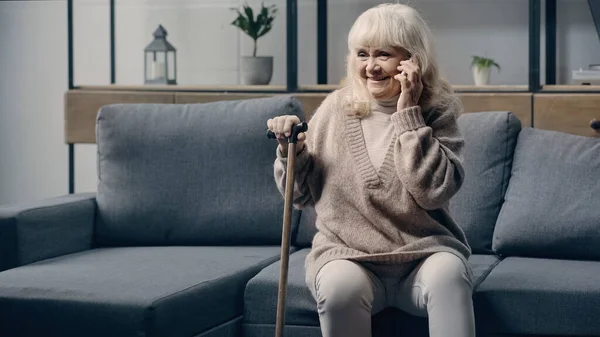 一位快乐的老妇人 她拿着手杖在客厅里用智能手机交谈 — 图库照片