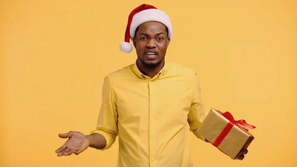 confused african american man in santa hat holding present and showing shrug gesture isolated on yellow