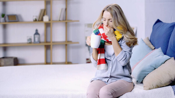 sick woman in colorful scarf drinking tea and coughing while sitting on bed at home