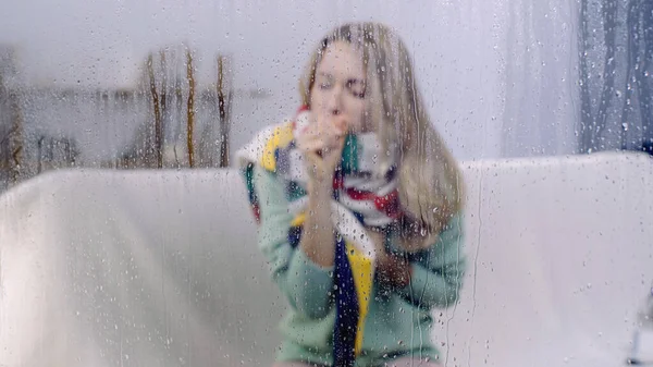 sick woman coughing behind wet window with rain drops