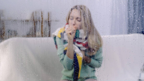 sick woman coughing behind wet window with rain drops 
