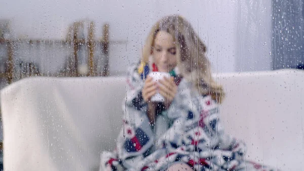生病的女人坐在五颜六色的毛毯下 在潮湿的窗户后面拿着一杯茶 还有雨滴 — 图库照片