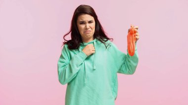 young and disgusted woman in turquoise hoodie holding stinky socks isolated on pink  clipart