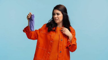 young and disgusted woman in orange denim jacket holding stinky socks isolated on blue  clipart