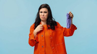 disgusted young woman in orange jacket holding stinky socks isolated on blue clipart