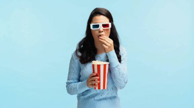 Üç boyutlu gözlüklü esmer bir kadın patlamış mısır yiyor ve mavi üzerine izlenmiş bir film izliyor.