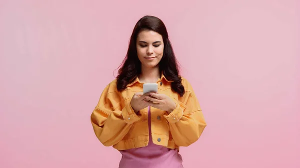 穿着橙色夹克笑着的年轻女人用智能手机发短信用粉色隔离 — 图库照片
