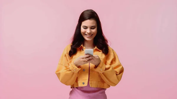 穿着橙色斜纹棉布夹克的快乐的年轻女人用智能手机发短信 用粉色隔开 — 图库照片