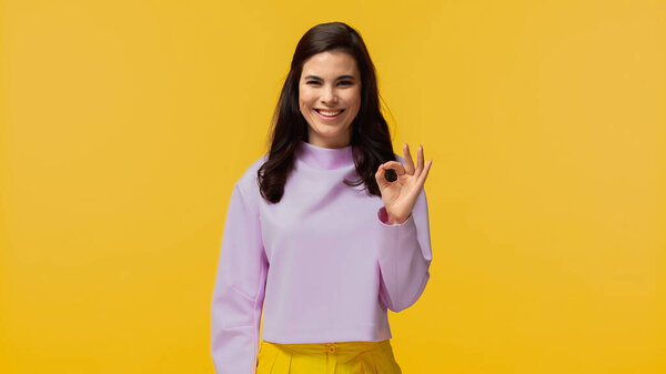 веселая молодая женщина в фиолетовой толстовке, зная хорошо и глядя на камеру, изолированную на желтый
