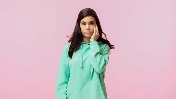 dissatisfied woman in turquoise hoodie looking at camera isolated on pink