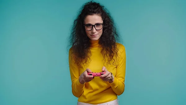 KYIV, UKRAINE - OCTOBER 1, 2021: brunette young woman in glasses holding joystick and playing isolated on blue — Stock Photo